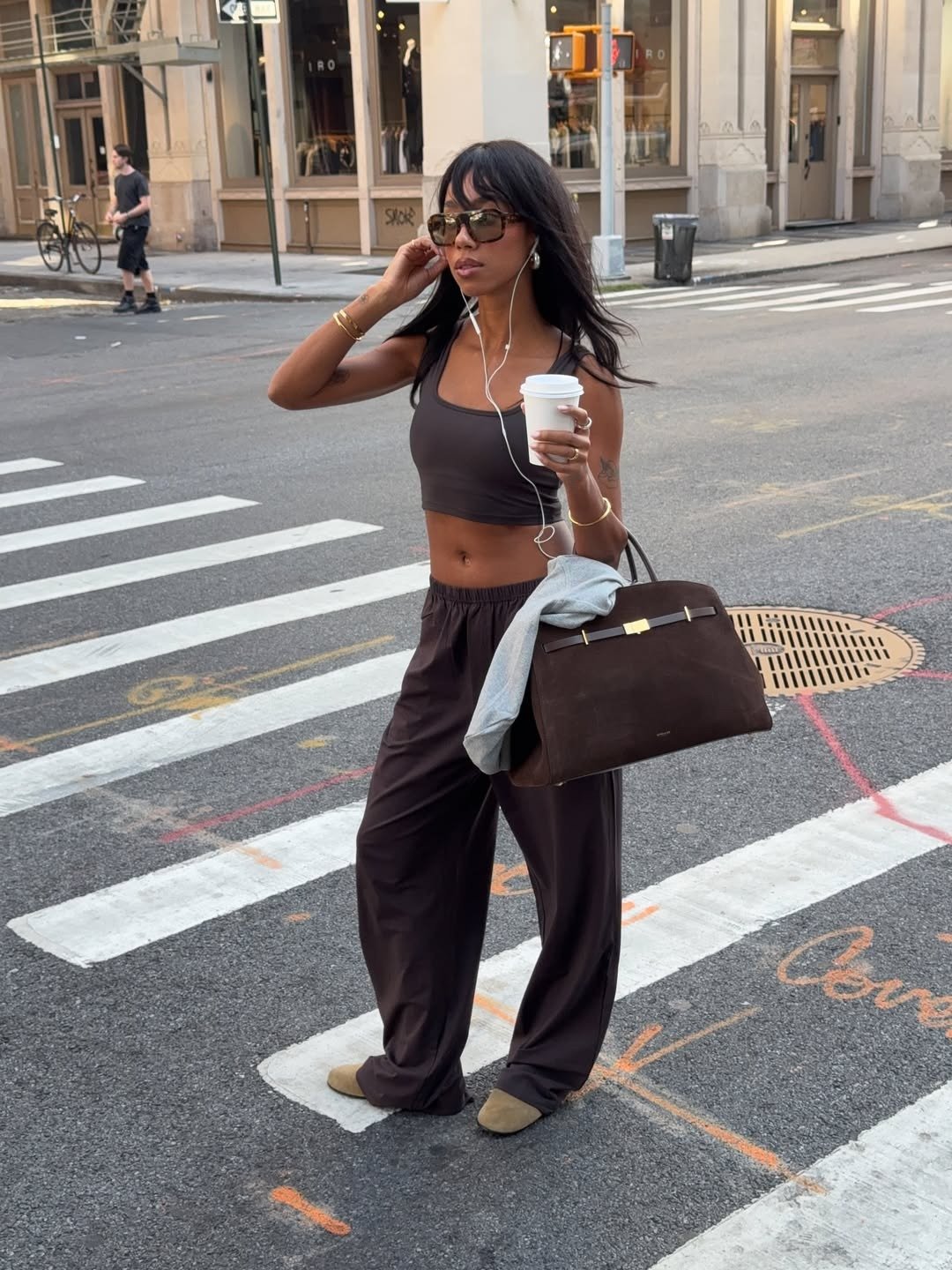 Modelo posa na rua com regatas cropped marrom de tecido leve, calça ampla combinando, bolsa grande e acessórios dourados.