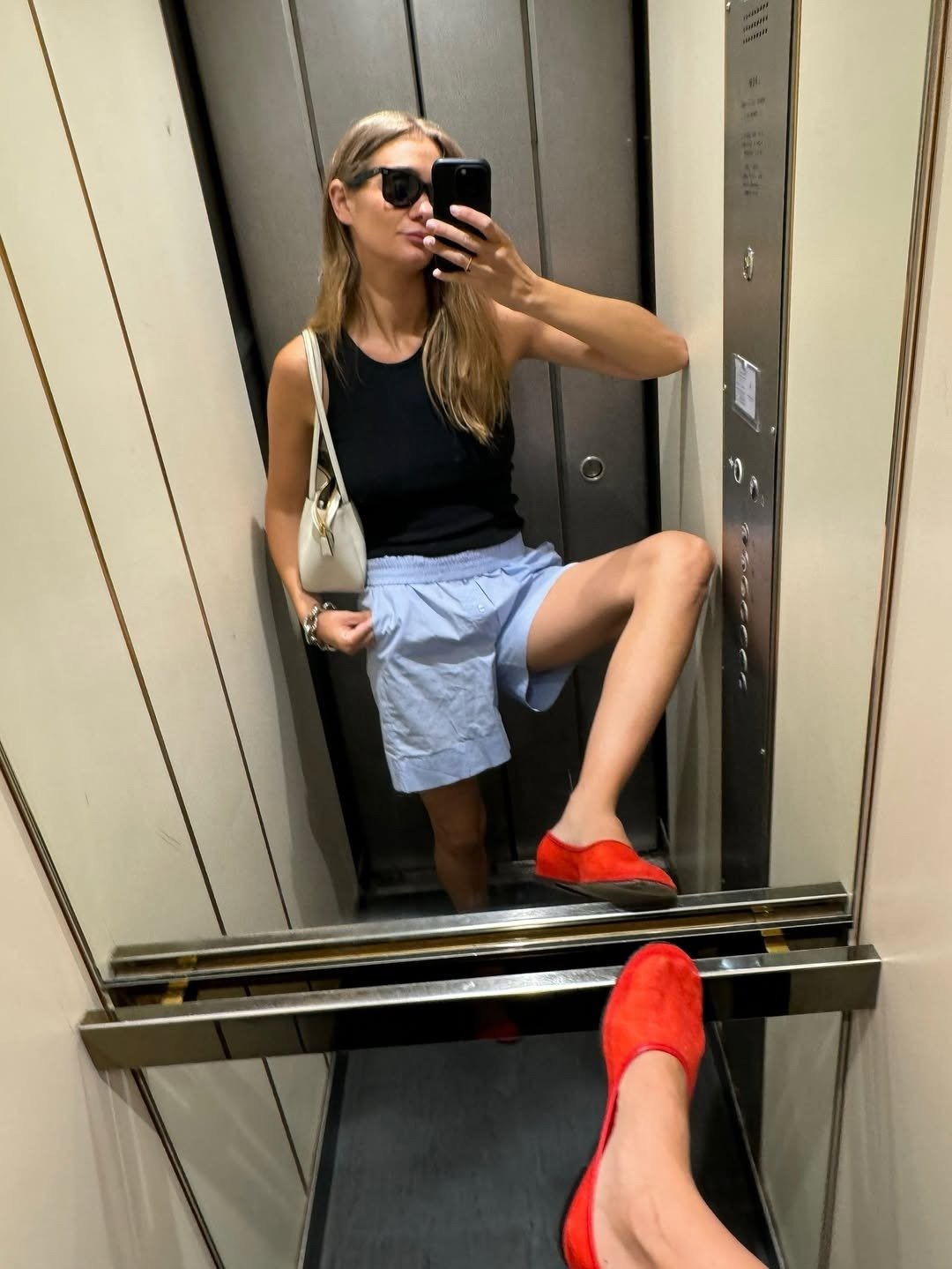 Modelo posa no elevador com short soltinho azul claro de tecido leve, regata preta justa e mocassim vermelho vibrante,