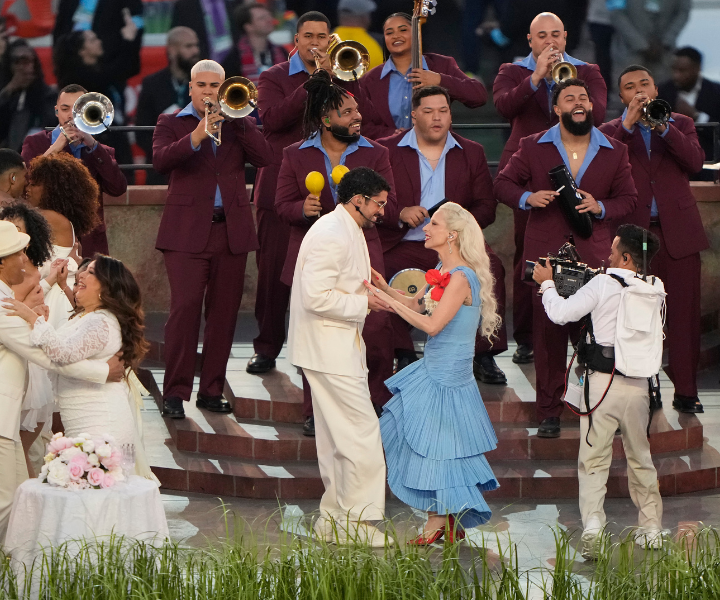 Bad Bunny veste terno branco clássico enquanto dança com Lady Gaga de vestido azul plissado e flores vermelhas.