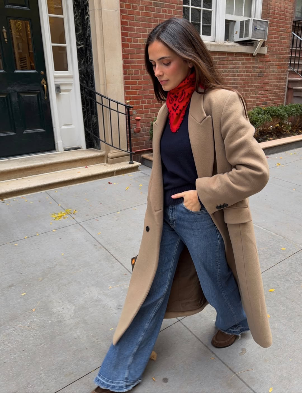Modelo caminha com bandana no pescoço vermelha estampada, casaco bege alongado, suéter preto e jeans flare azul.
