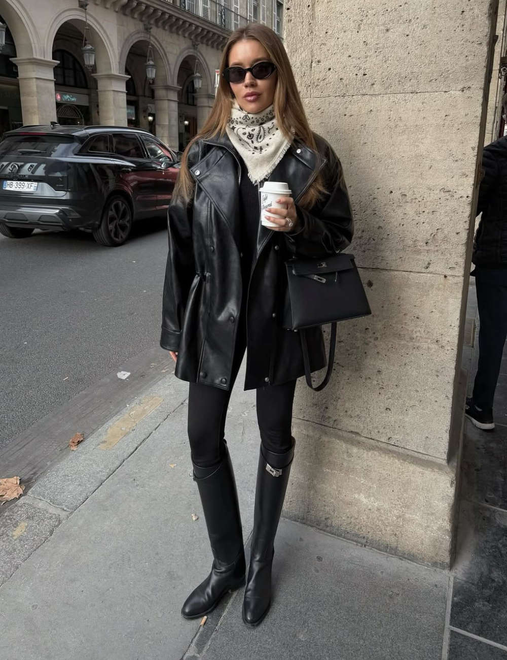 Modelo posa com bandana no pescoço branca estampada, jaqueta oversized de couro preta, legging e botas longas.