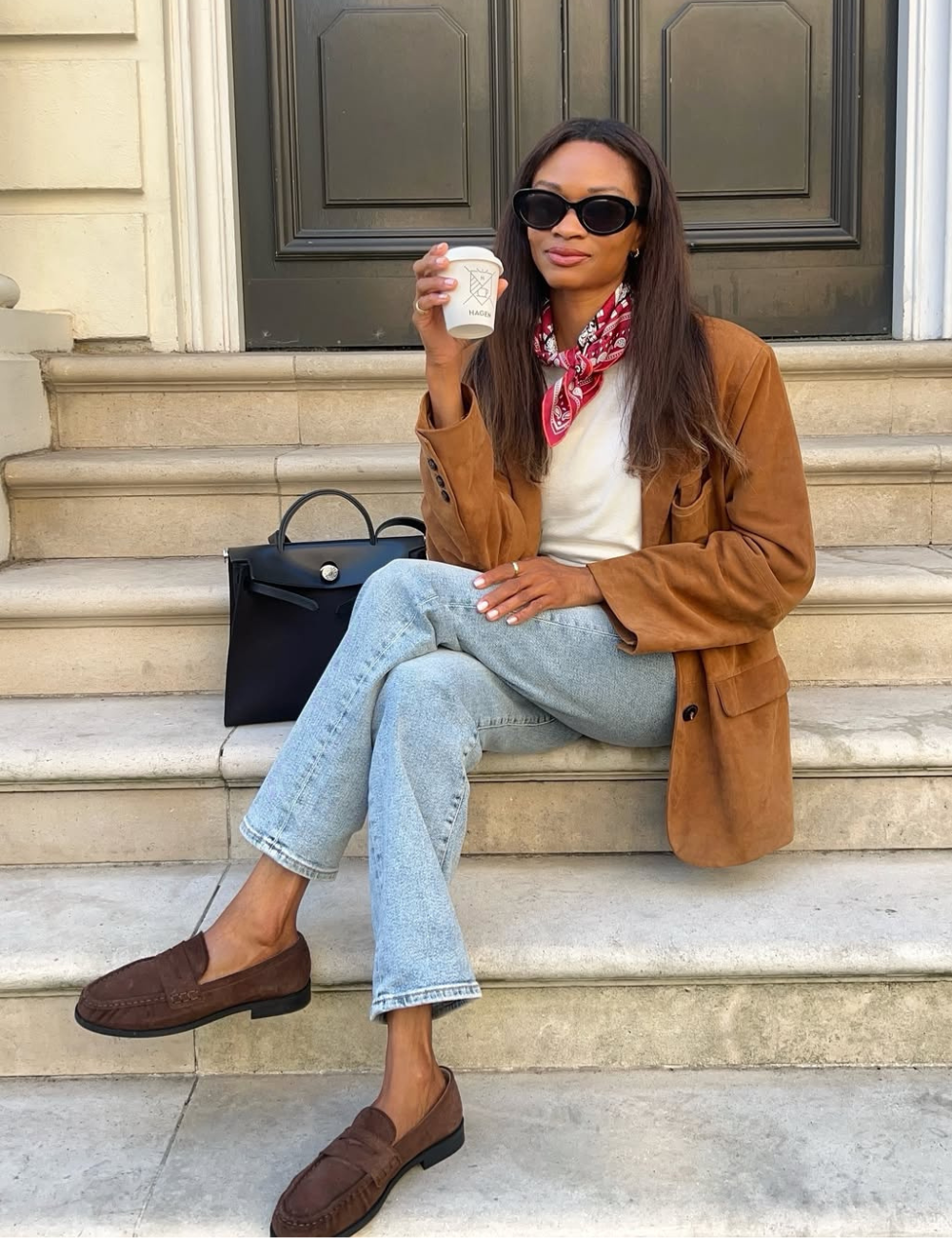 Mulher sentada com bandana no pescoço vermelha estampada, blazer caramelo, jeans claro e mocassim marrom.