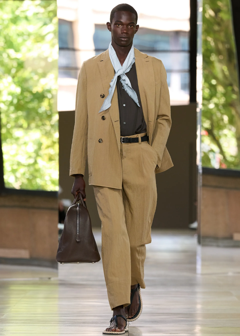 Modelo caminha com postura confiante, usando bandana no pescoço azul clara, terno bege de linho, camisa preta e bolsa marrom.