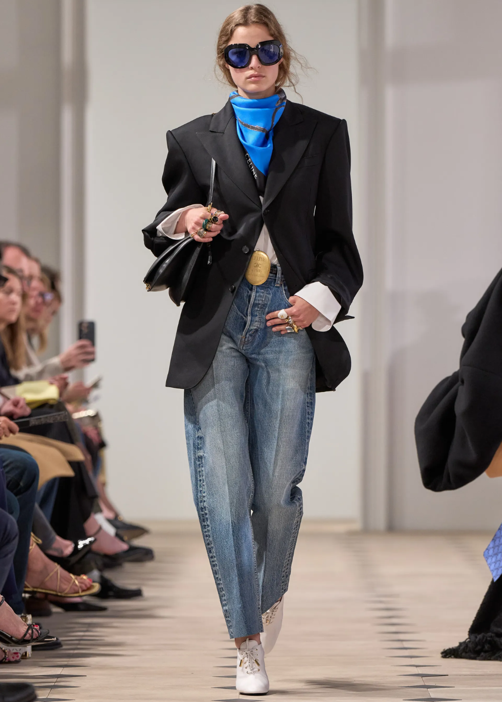 Modelo caminha com bandana no pescoço azul vibrante, blazer preto oversized, jeans claro e acessórios dourados marcantes.