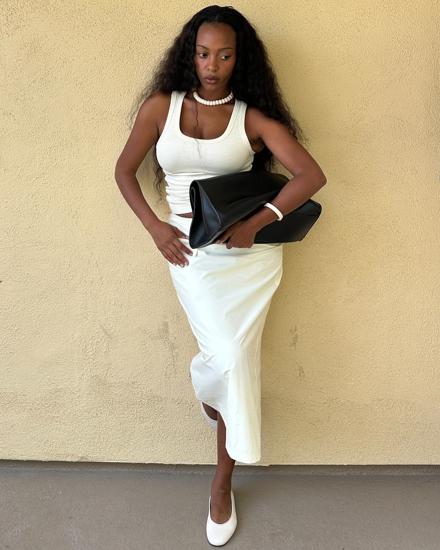 Modelo posa com postura confiante segurando bolsas elegantes preta de couro estruturado, combinando com look branco