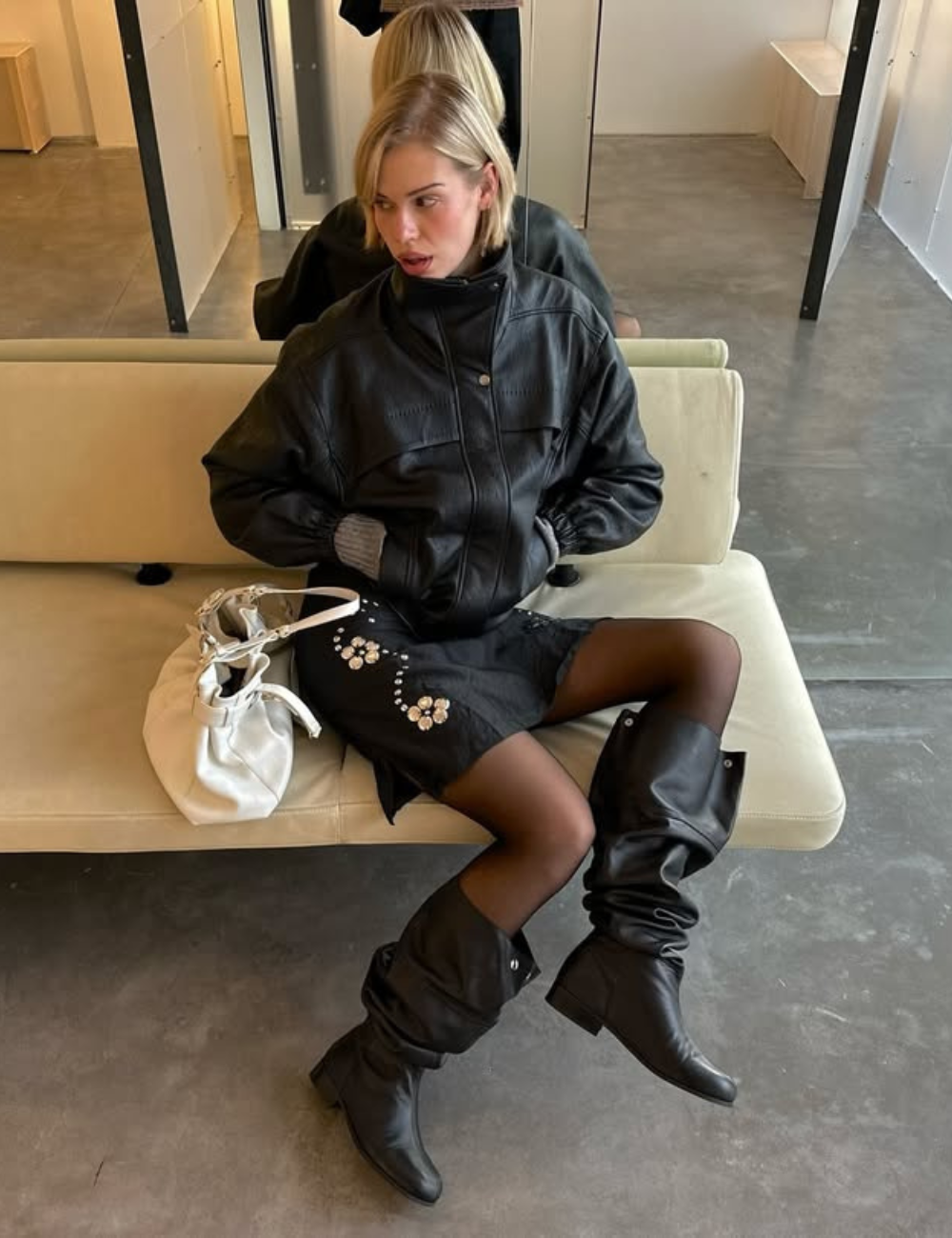 Modelo senta com postura confiante usando bota slouchy de couro preta, saia preta com bordados e jaqueta oversized preta.