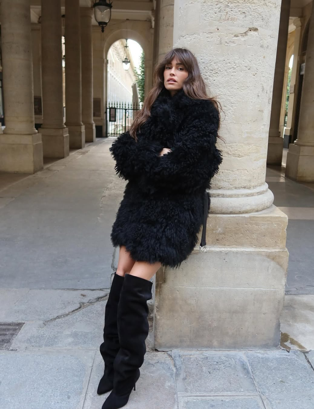 Modelo posa encostada com casaco preto peludo e bota slouchy preta de camurça, combinando elegância e conforto urbano.