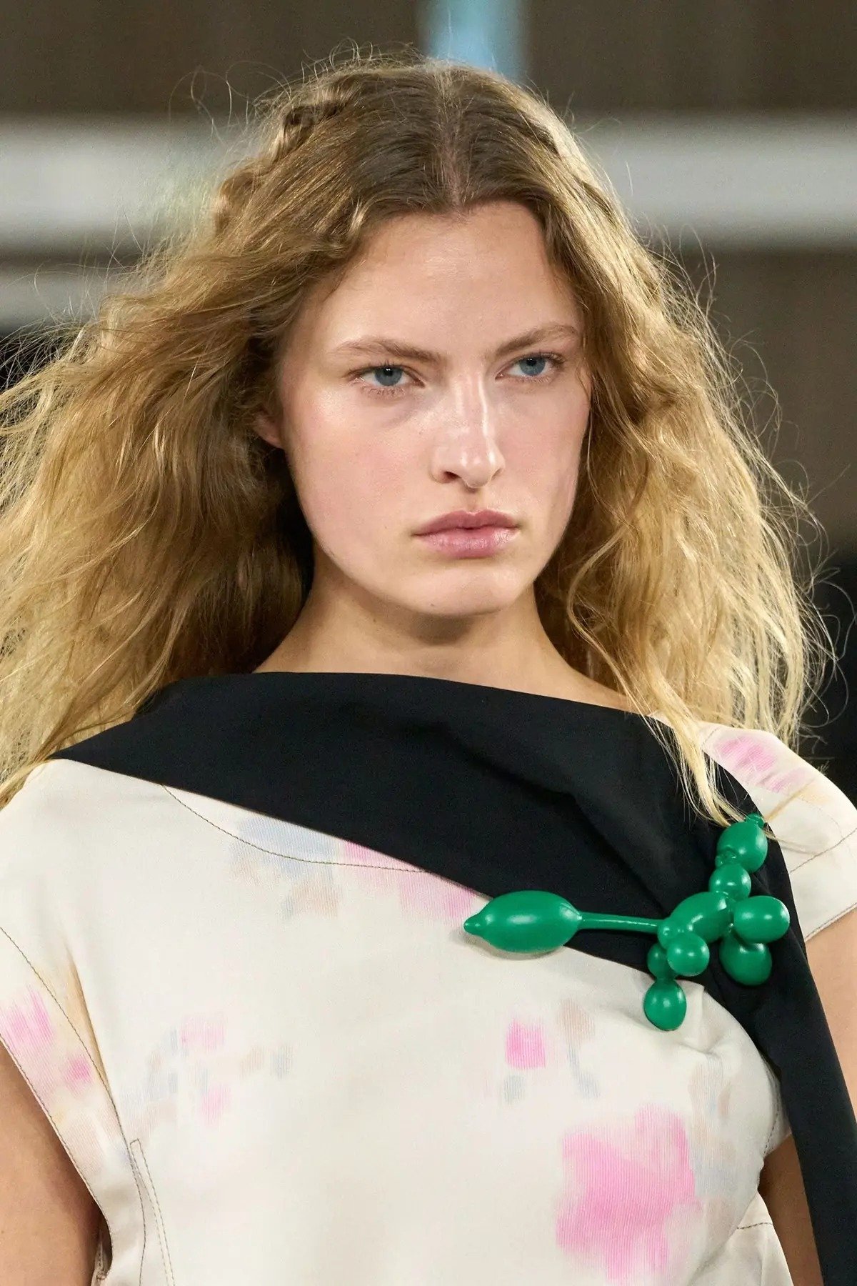 Modelo com expressão séria veste blusa off-white com detalhes rosa e preto, destacando broche verde em formato de balão