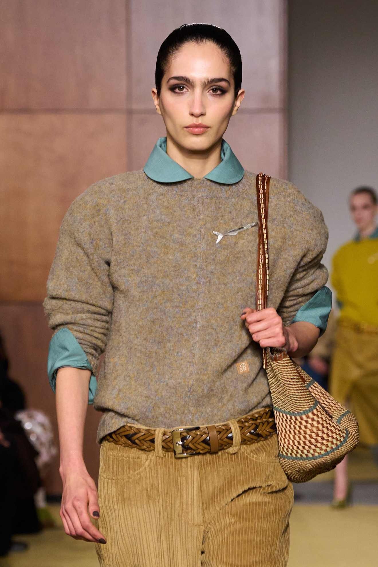 Modelo caminha com postura firme usando suéter cinza com gola azul, broche prateado em formato de peixe e calça mostarda de