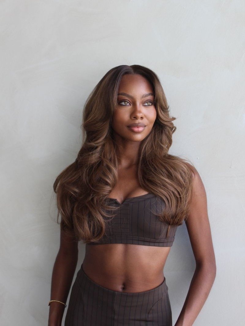 Modelo posa com cabelo marrom avelã longo e ondulado, usando top cropped e saia marrom com listras finas, em clima elegante