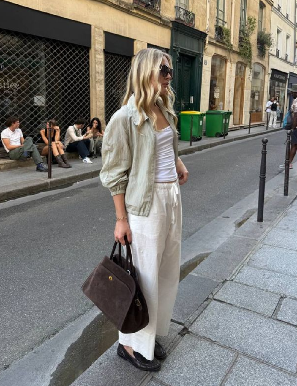 Modelo posa na rua com looks com calça de linho cru ampla, regata branca justa, jaqueta leve bege e bolsa marrom.