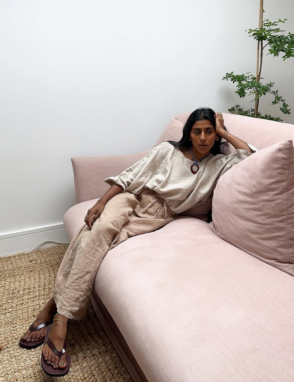 Modelo relaxa no sofá rosa usando look com calça de linho cru de modelagem ampla, blusa de algodão bege e sandálias