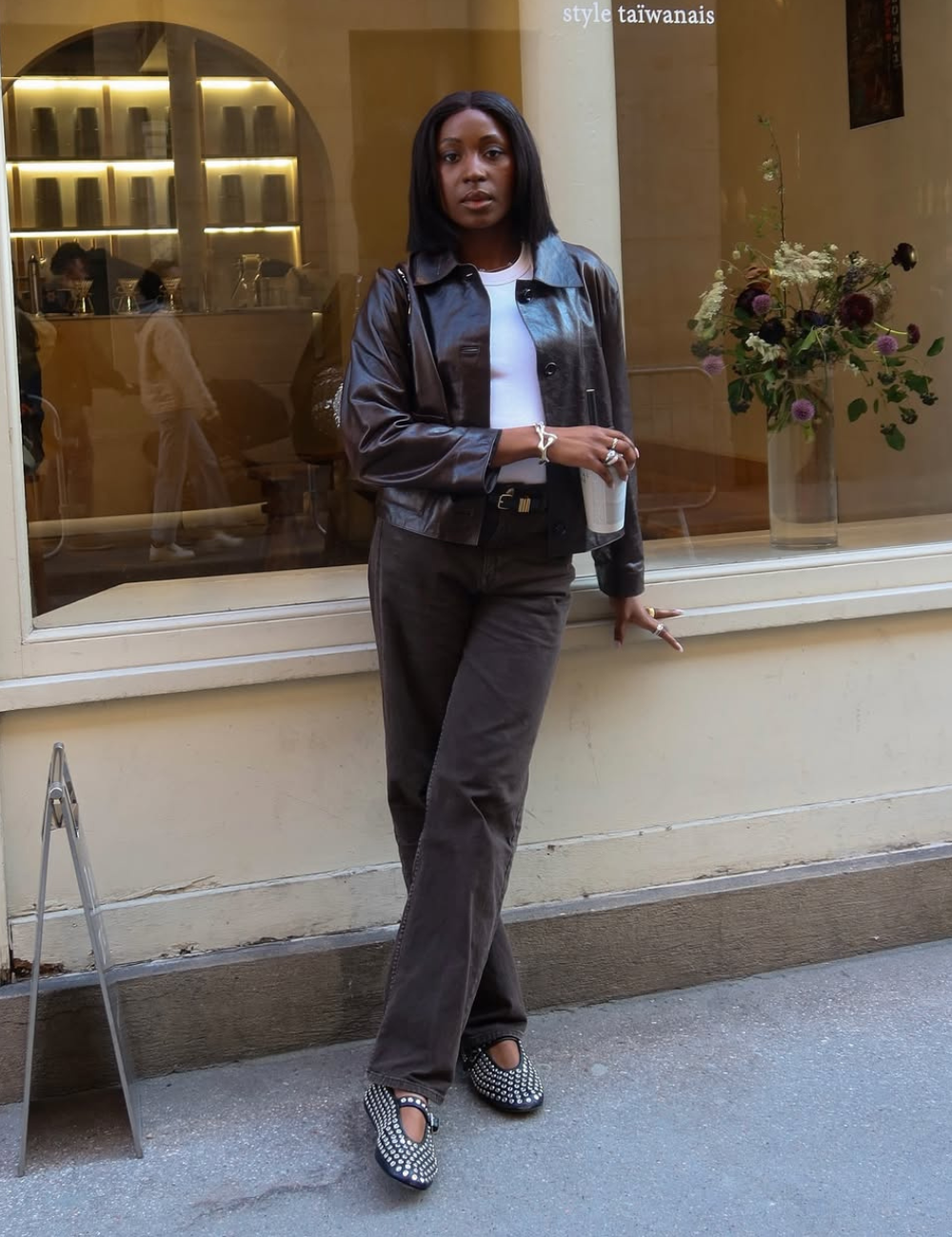 Modelo de calça jeans elegante em tom escuro com corte reto, combinada a jaqueta de couro marrom e blusa branca.