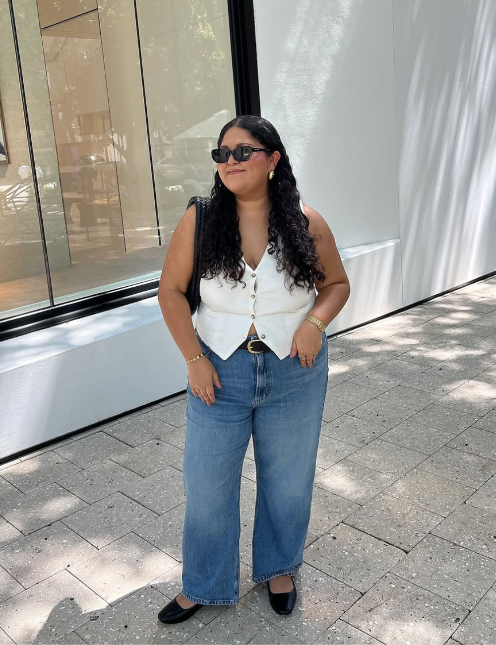 Modelo de calça jeans elegante em lavagem média e corte reto, combinada com colete branco estruturado e acessórios dourados.