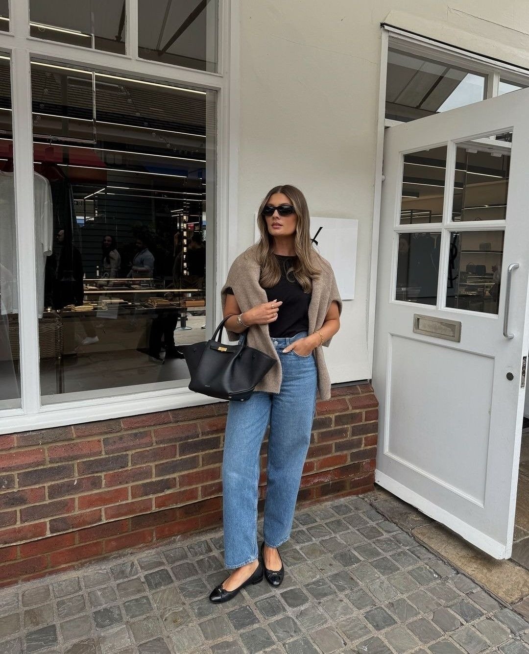 Modelo posa com calça mom jeans azul clara de algodão, camiseta preta, cardigan bege sobre os ombros e bolsa preta