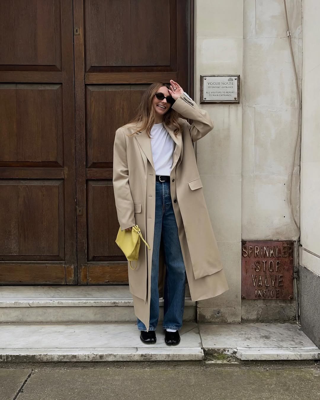 Modelo posa sorrindo com calça mom jeans azul de lavagem média, camiseta branca e sobretudo bege longo