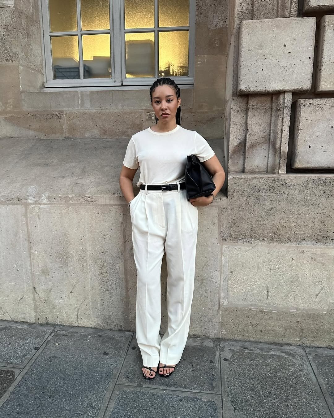 Modelo posa com calça social feminina de alfaiataria branca, camiseta básica, cinto preto e bolsa estruturada, em clima