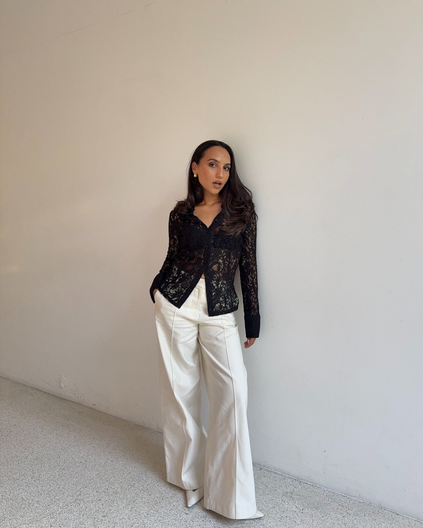 Modelo posa com calça social feminina branca de modelagem ampla e tecido estruturado, combinada a camisa preta rendada, em