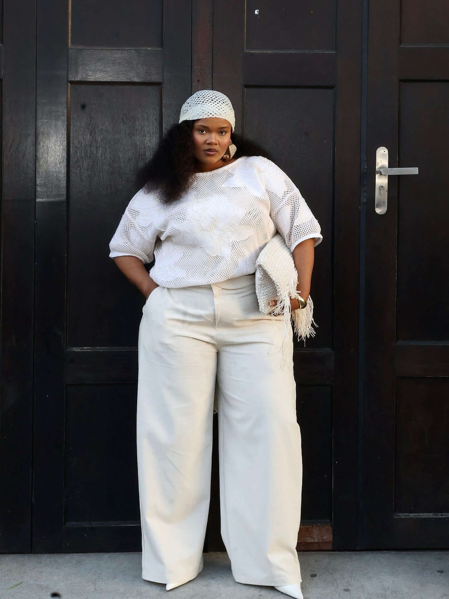 Modelo posa com calça social feminina de alfaiataria ampla em tom off-white, combinada a blusa de tricô vazada e acessórios