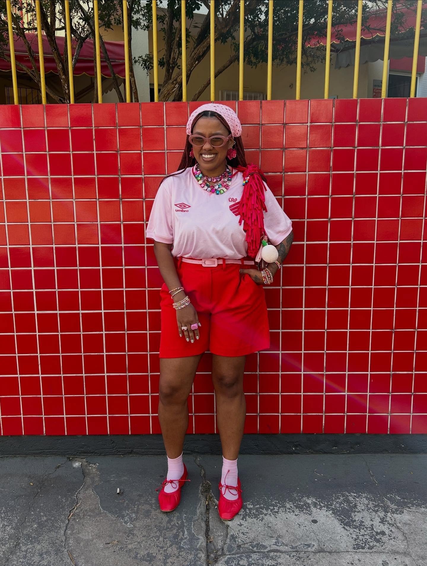 Modelo posa sorridente com camisa de time bonita rosa clara de malha leve, combinada a shorts vermelhos e acessórios