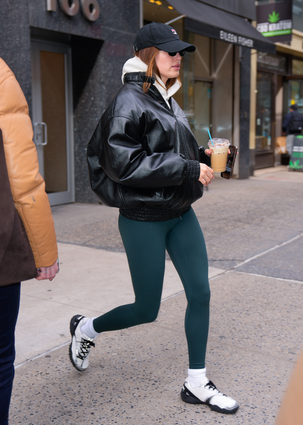 Modelo caminha com tênis da moda de 2026 em branco e preto, combinando jaqueta oversized preta de couro e legging verde.