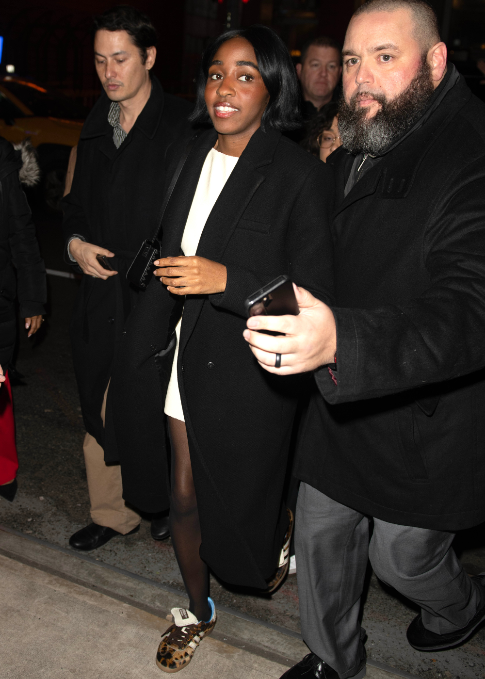 Mulher caminha confiante com vestido branco, sobretudo preto e tênis da moda de 2026 em animal print com detalhes azuis.