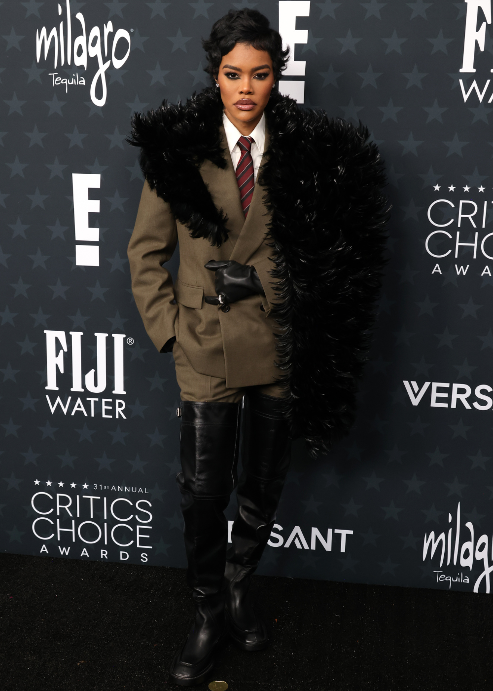 Modelo posa com blazer oversized cáqui, gravata listrada, bota over the knee e maxi casaco de pele preta.