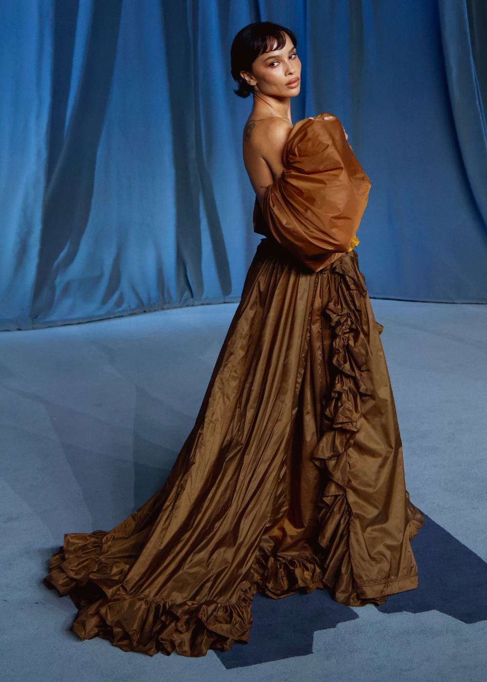 Modelo posa de costas e olhar sobre o ombro com vestido longo marrom em tecido leve e volumoso.