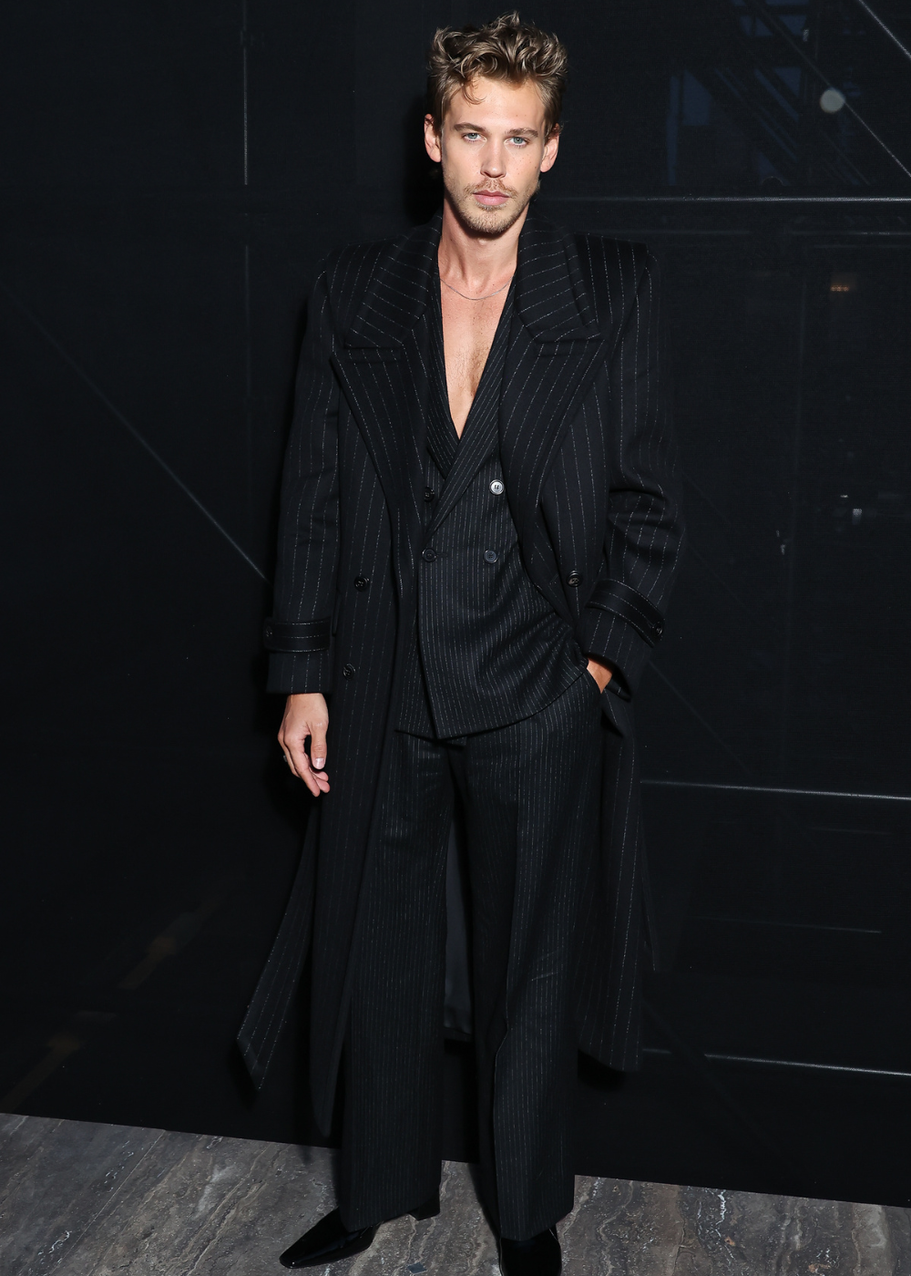 Modelo posa com elegância usando conjunto de alfaiataria preta risca de giz, casaco longo estruturado e calça ampla.