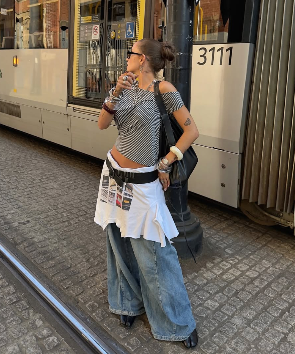 Modelo posa de lado na rua com blusa listrada ombro a ombro, jeans largo, saia branca sobreposta e cinto Y2K preto.