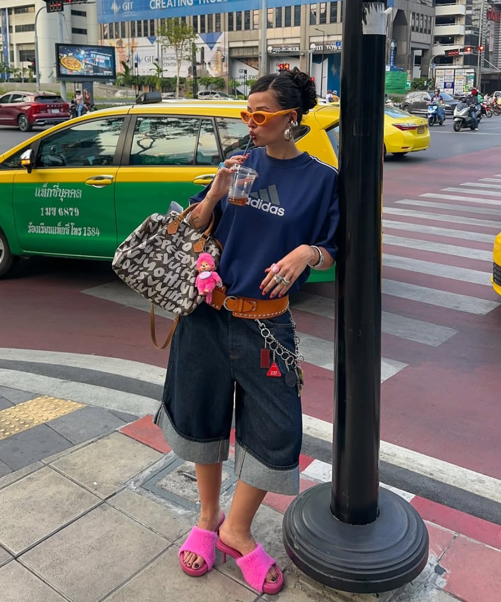 Modelo posa na rua com cinto Y2K caramelo de couro cravejado, bermuda jeans larga, camiseta azul e sandálias rosa peludas.