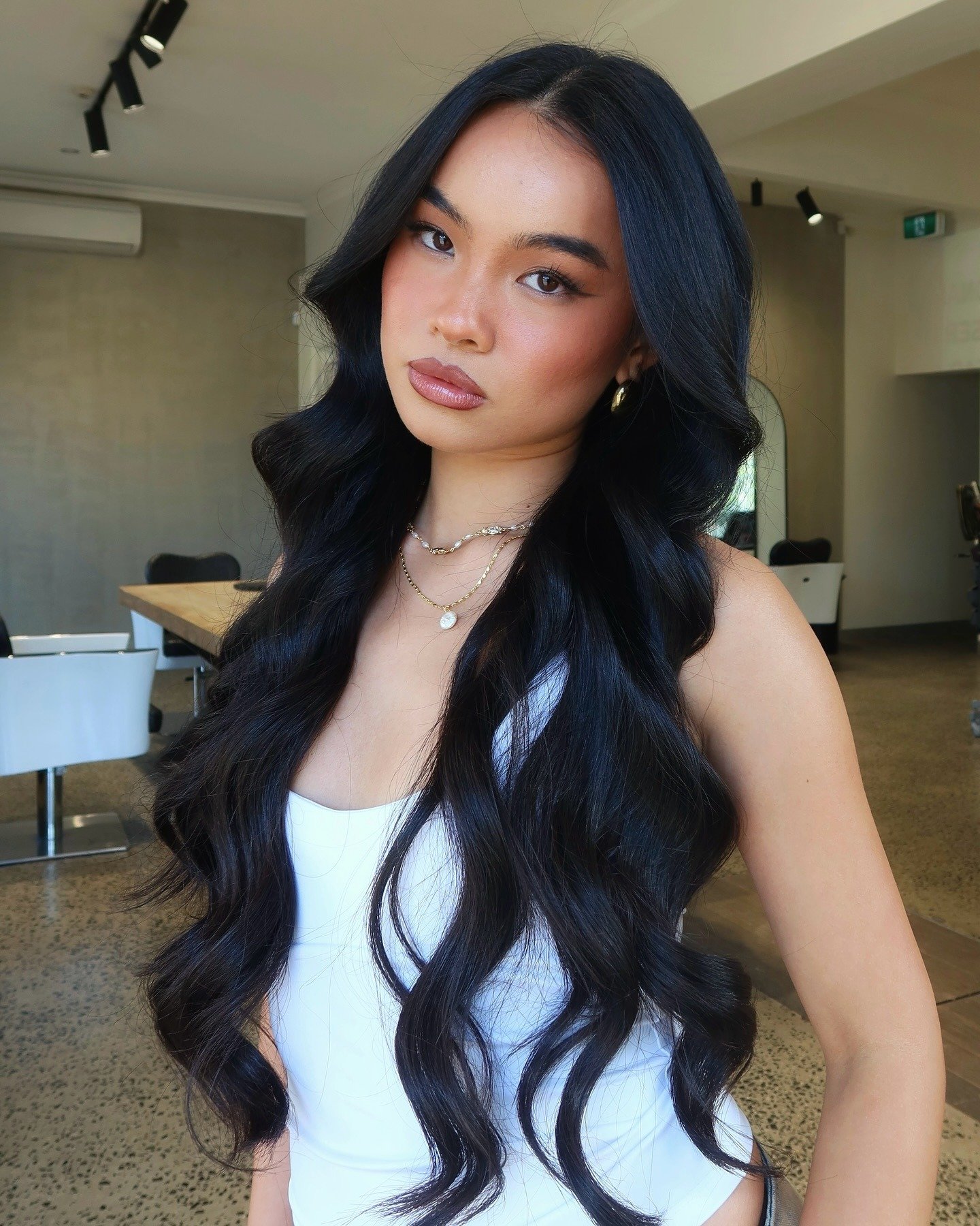 Modelo posa com expressão confiante de como fazer babyliss em cabelos longos e pretos, usando regata branca