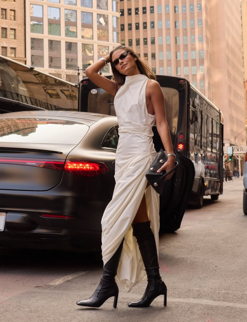 Modelo posa confiante na rua com vestido branco drapeado de tecido fluido e bota preta de cano alto e salto fino.