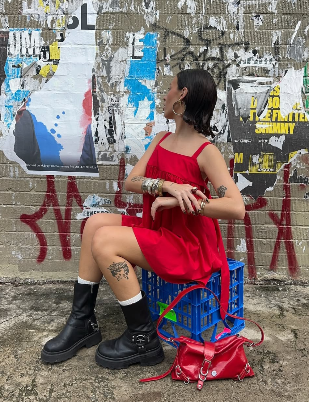 Modelo sentada em caixa azul usa vestido vermelho solto de alças finas com botas pretas robustas.