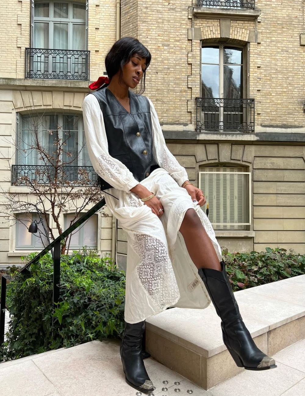 Modelo senta com vestido branco longo de renda e colete preto de couro, combinando com bota preta de cano alto.