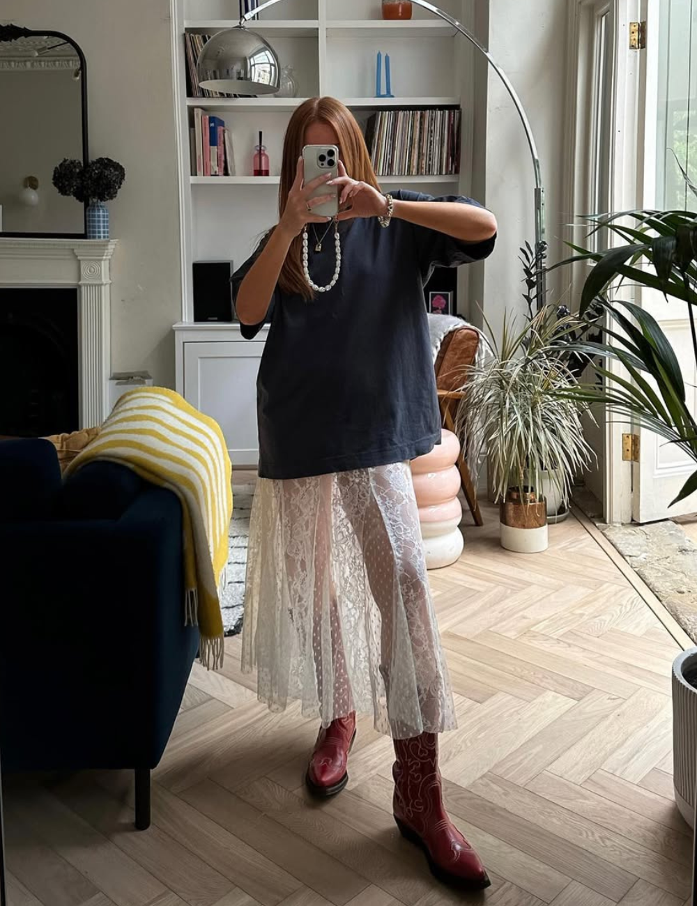 Modelo posa segurando celular com vestido branco de renda transparente e camiseta preta oversized, combinando bota vermelha.