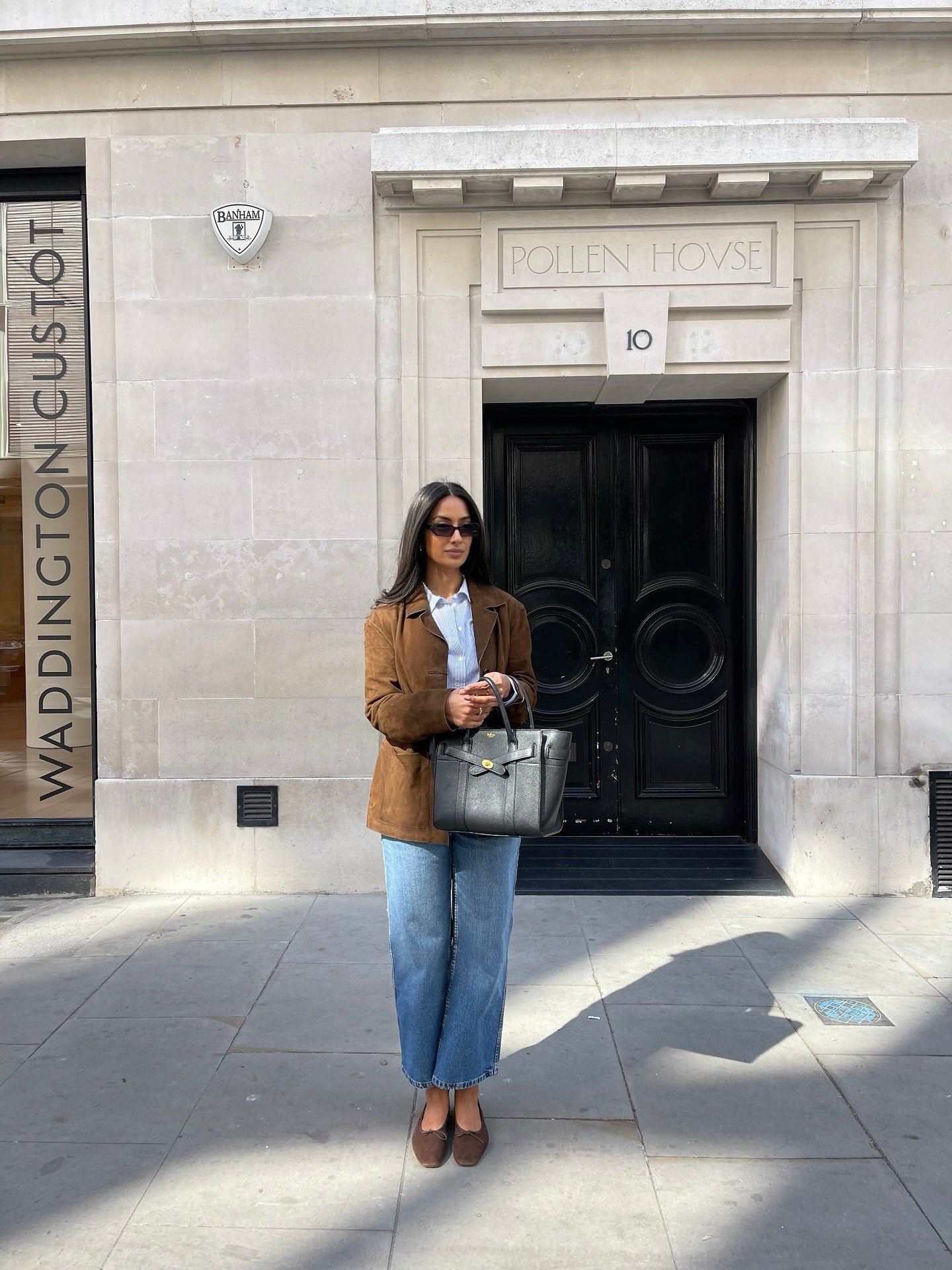Mulher posa com camisa branca listrada, calça jeans reta azul clara e blazer marrom em camurça, criando look casual