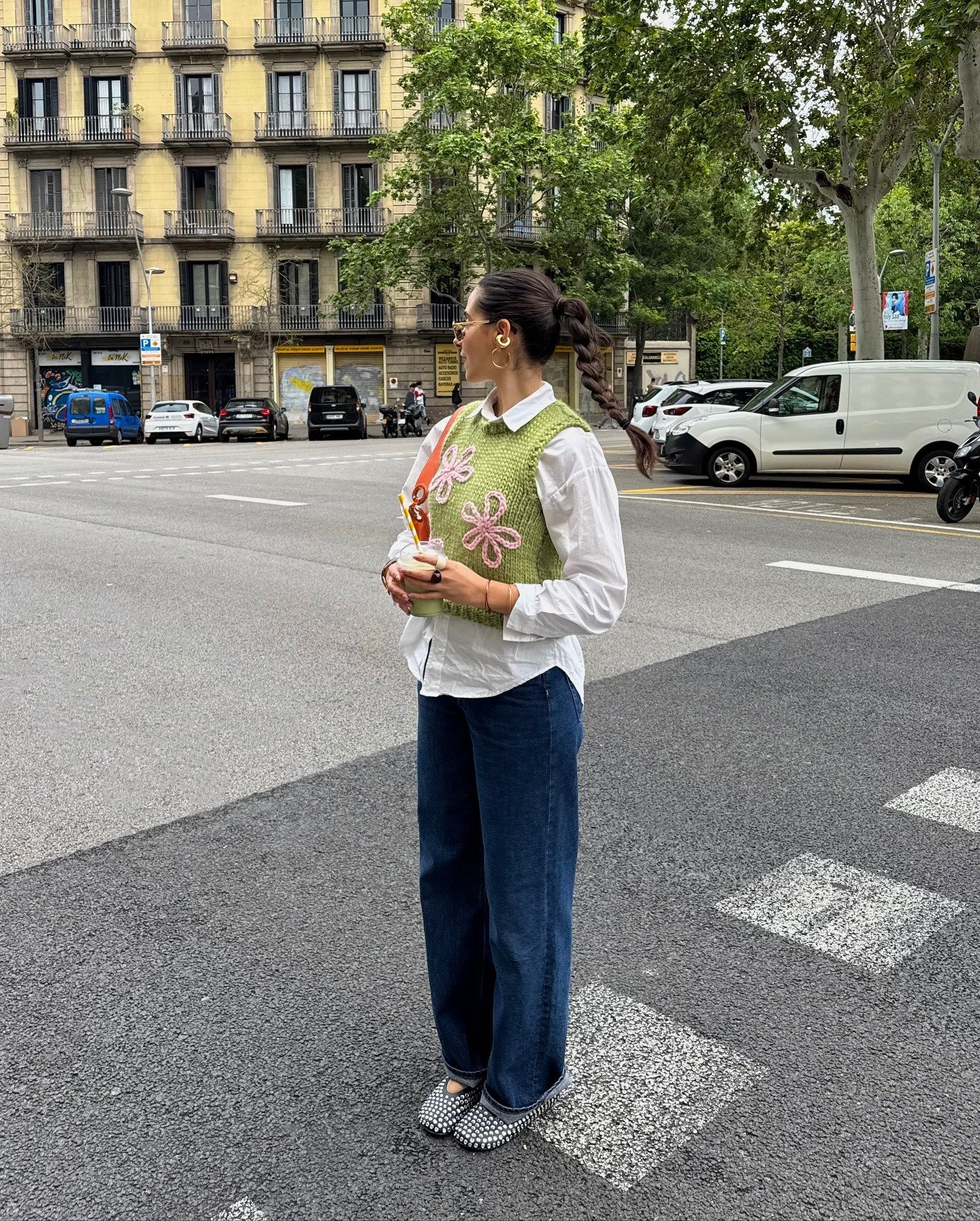 Modelo posa na rua com camisa branca de algodão, colete verde tricô com flores rosas e calça jeans reta azul escuro