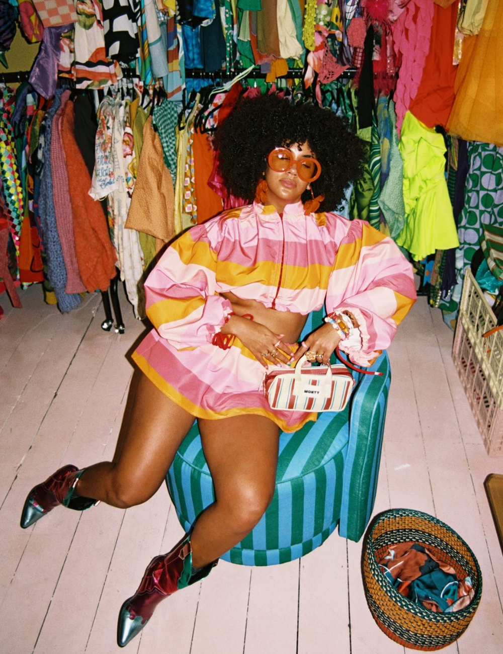 Mulher posa sentada em poltrona azul com look cropped e saia rosa e amarelo em tecido leve, celebrando consumo sustentável.