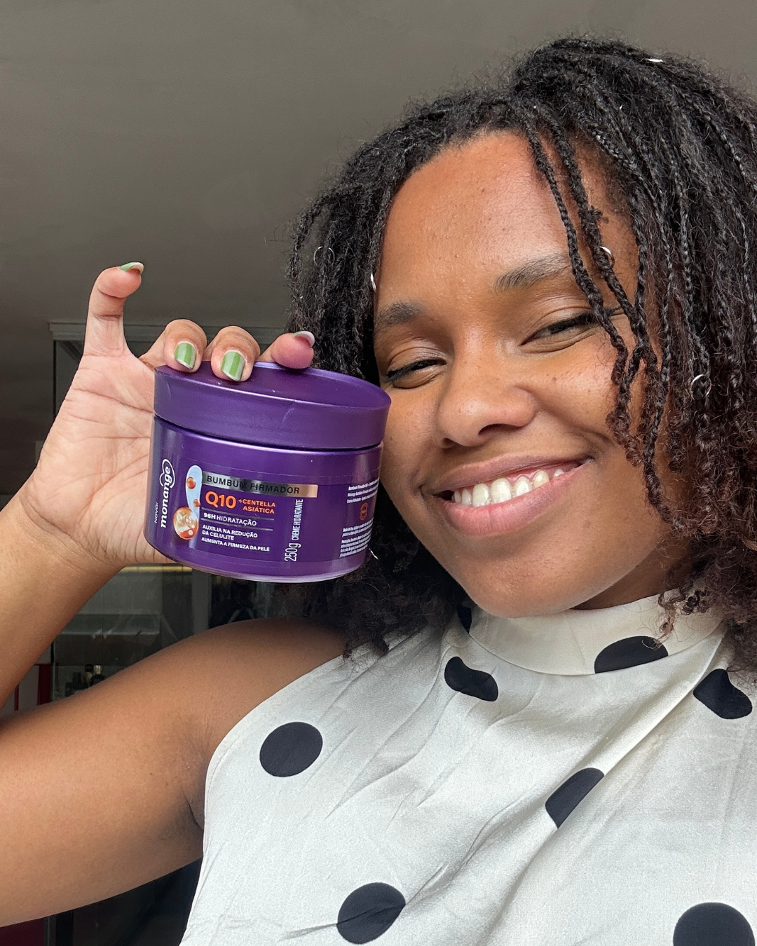 Mulher sorridente segura pote roxo de creme para celulite, vestindo blusa branca com bolinhas pretas, em ambiente interno