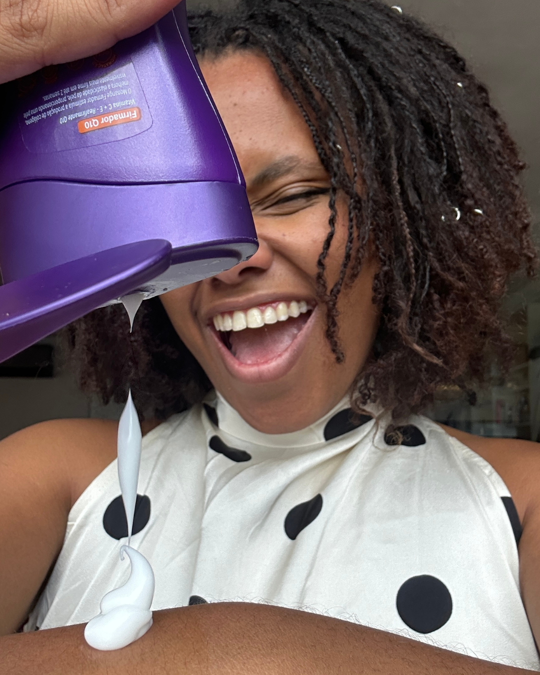 Mulher sorridente aplica creme para celulite branco, com embalagem roxa, vestindo blusa branca de bolinhas pretas