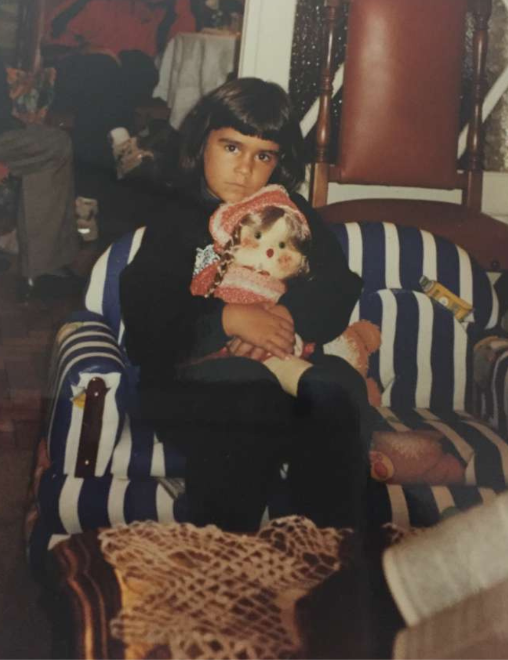 Criança de cabelos curtos e escuros veste moletom preto, senta em poltrona listrada azul e branca abraçando boneca.