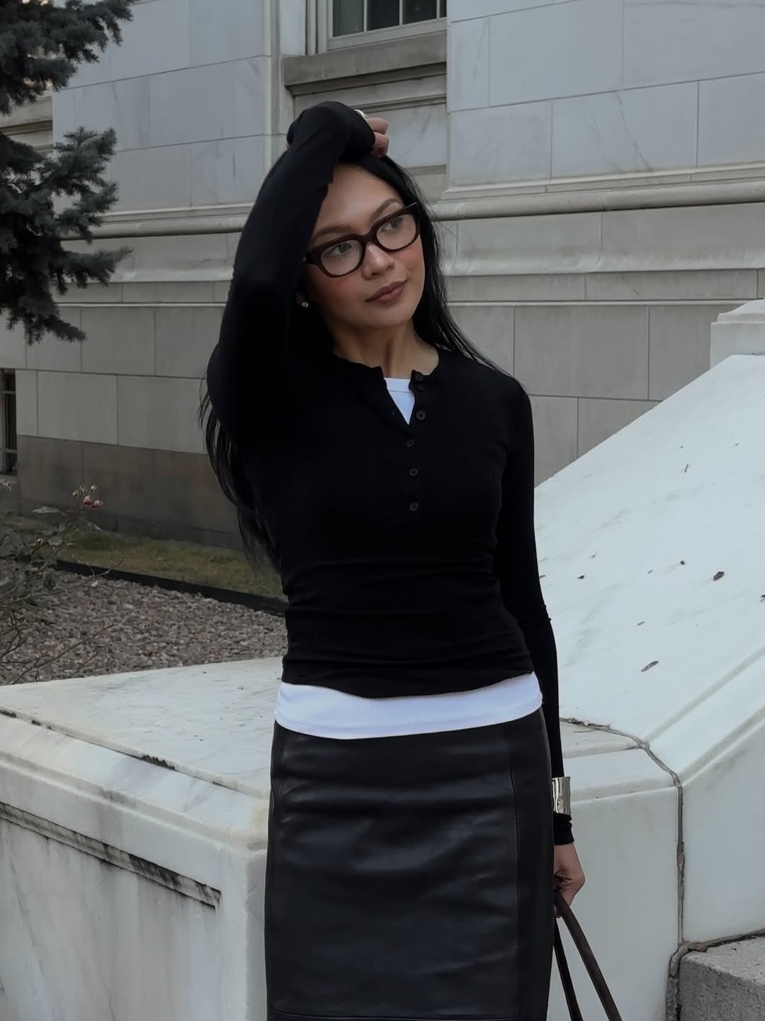 Modelo posa com tricô leve preto de botões, sobreposição branca e saia de couro preta, em clima urbano e sofisticado.