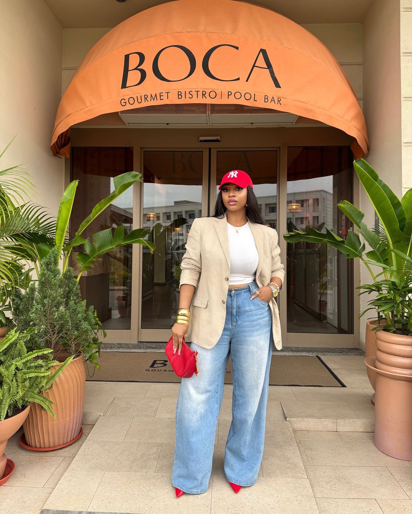 Mulher posa em estilo casual chique com blazer bege, cropped branco, jeans wide leg, acessórios dourados e boné vermelho