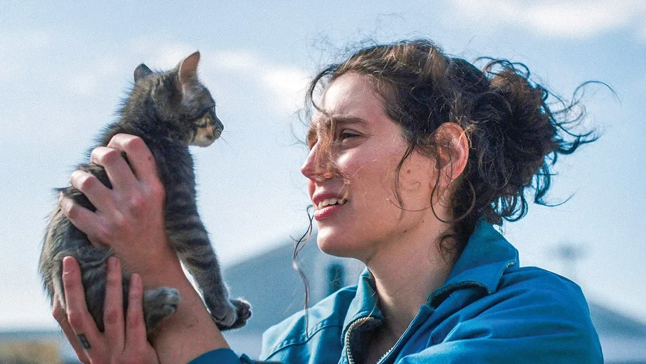 Eva Victor segura um gatinho cinza de pelagem macia, veste jaqueta azul de tecido resistente e expressa ternura sob luz