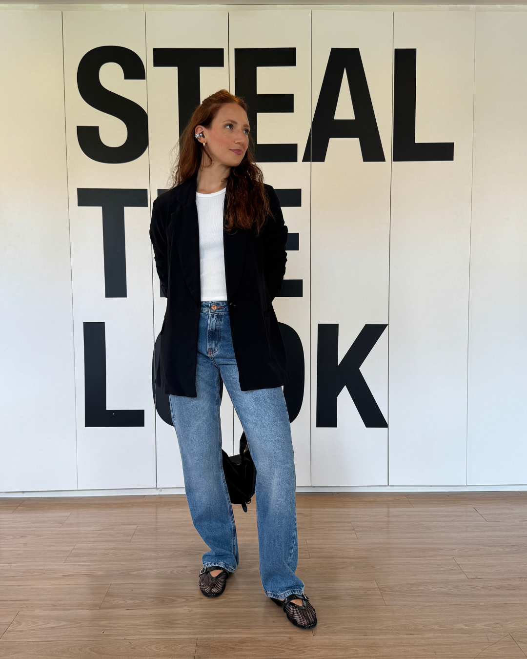 Mulher com fone de ouvido sem fio preto, blazer alongado, camiseta branca e jeans azul em pose descontraída diante da parede