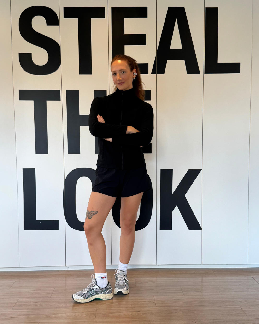 Modelo posa com fone de ouvido sem fio preto, jaqueta e shorts esportivos pretos, tênis cinza e ambiente minimalista branco