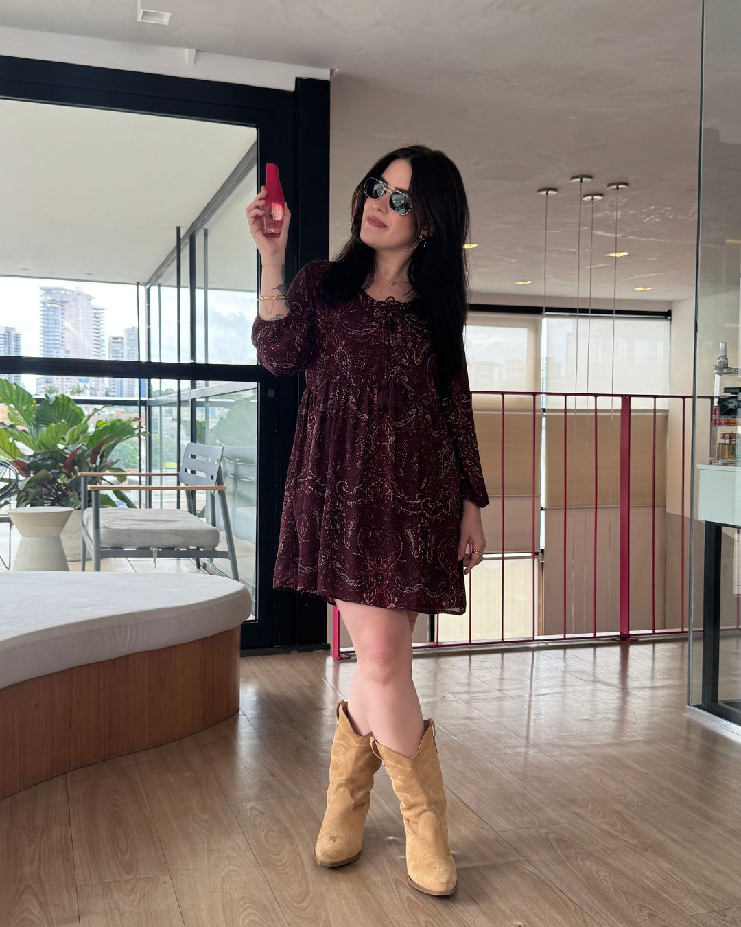 Modelo posa com vestido vinho estampado de modelagem solta e botas bege, segurando fragrâncias intensas em ambiente moderno