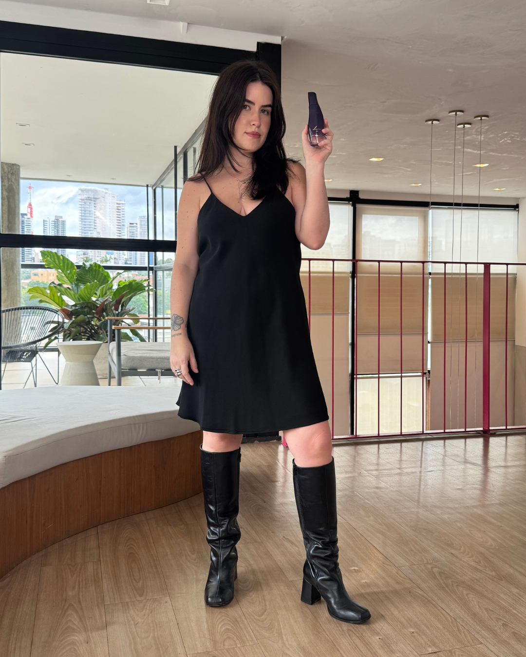 Modelo posa com vestido preto de alças finas e bota de couro preta, segurando frasco roxo que sugere fragrâncias intensas.