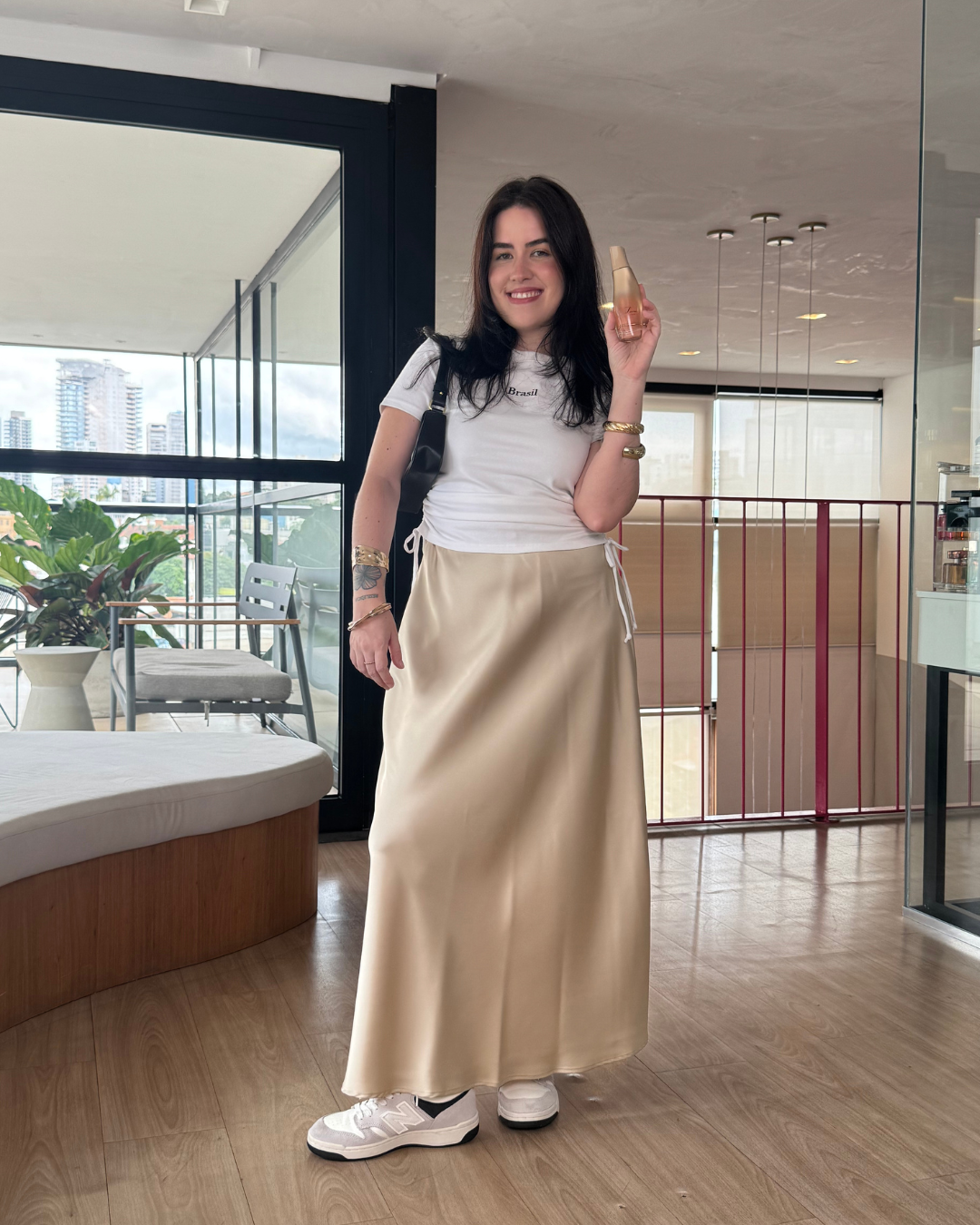Modelo sorri e posa segurando fragrâncias intensas, vestindo saia longa bege acetinada, camiseta branca justa e tênis casuais